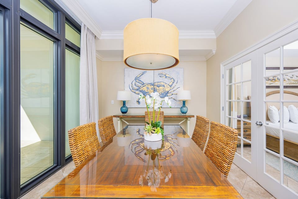 This dining area features a stylish table with woven chairs, a vibrant crab artwork, and a glimpse of the cozy bedroom beyond