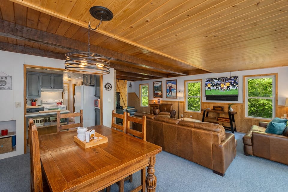 Relax in your cozy cabin retreat with warm wood ceilings, comfy seating, and big windows framing lush green views.