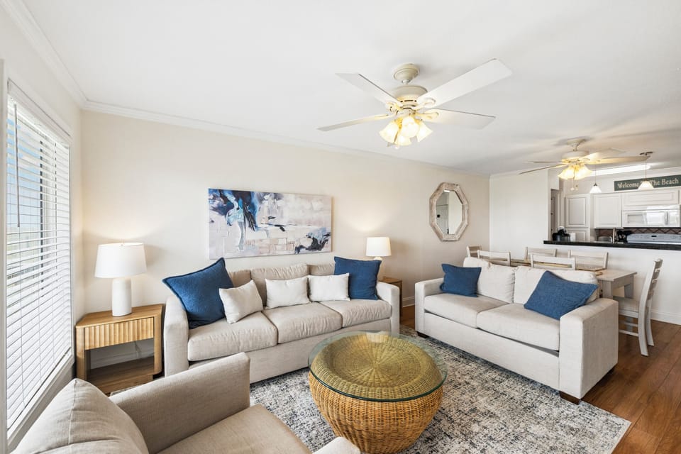Relax in your bright, coastal living room featuring plush seating, calming blue accents, a woven ottoman, and ceiling fans for comfort. The open layout flows seamlessly into the dining and kitchen area.