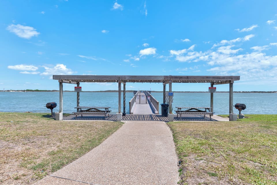 Waterfront pavilion with picnic tables and fishing pier extends into calm waters under blue skies.
