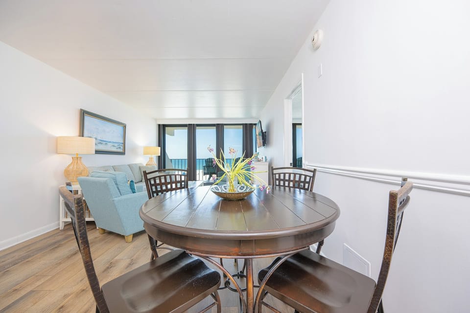 The living room features a round dining table, comfortable chairs, and a stunning beachfront view through large windows