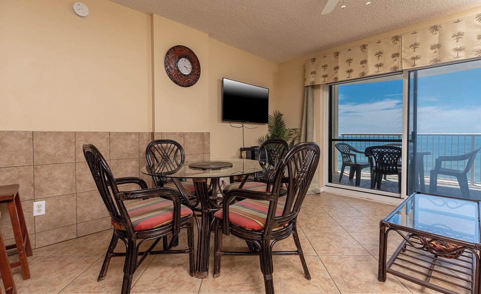 The dining area features a glass table, colorful seating, a TV, and a stunning beachfront view through sliding glass doors
