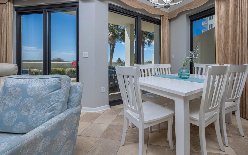 Bright dining area features a white table surrounded by chairs, with large windows offering a view of palm trees outside