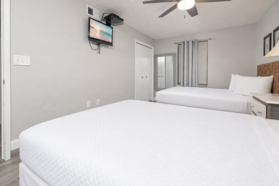 Guest room featuring two queen beds, a wall-mounted TV, and a large mirror, complemented by soft gray tones and natural light