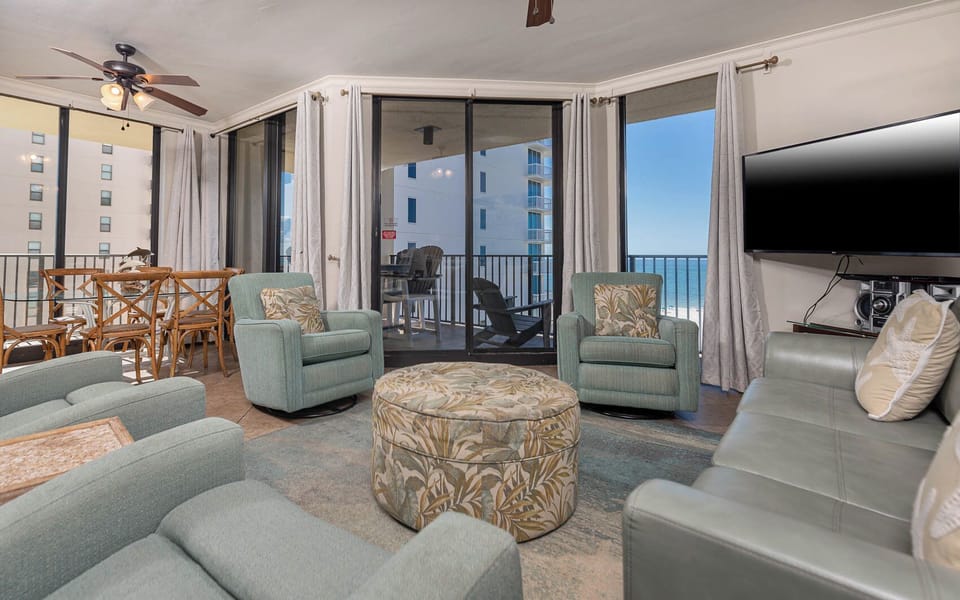 Cozy seating surrounds a patterned ottoman, with sliding glass doors revealing a balcony and a glimpse of the beach beyond
