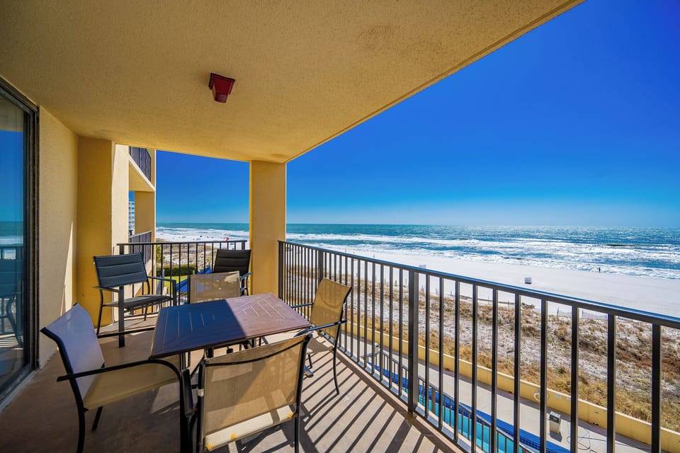The balcony features a table and chairs, offering a serene view of the beach and vibrant blue skies