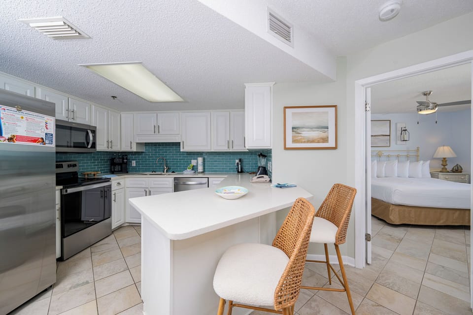 Bright kitchen area featuring white cabinetry, a breakfast bar with two woven stools, and a glimpse of the master bedroom