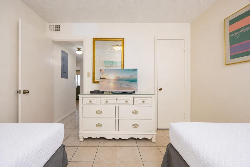 This guest bedroom features two beds, a TV on a stylish white dresser, and a welcoming view into the hallway