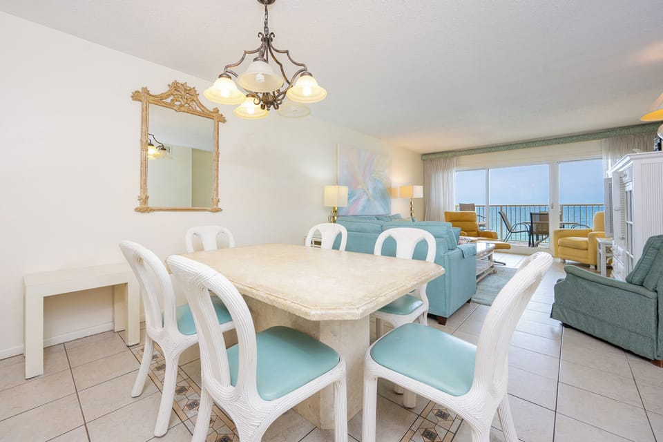 This dining area features a light stone table and white chairs, with a stunning beachfront view visible through large windows