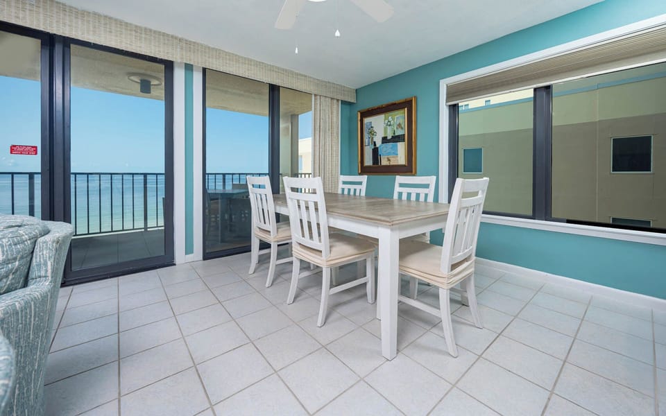This dining area features a light wood table surrounded by white chairs, with a breathtaking beach view through large windows