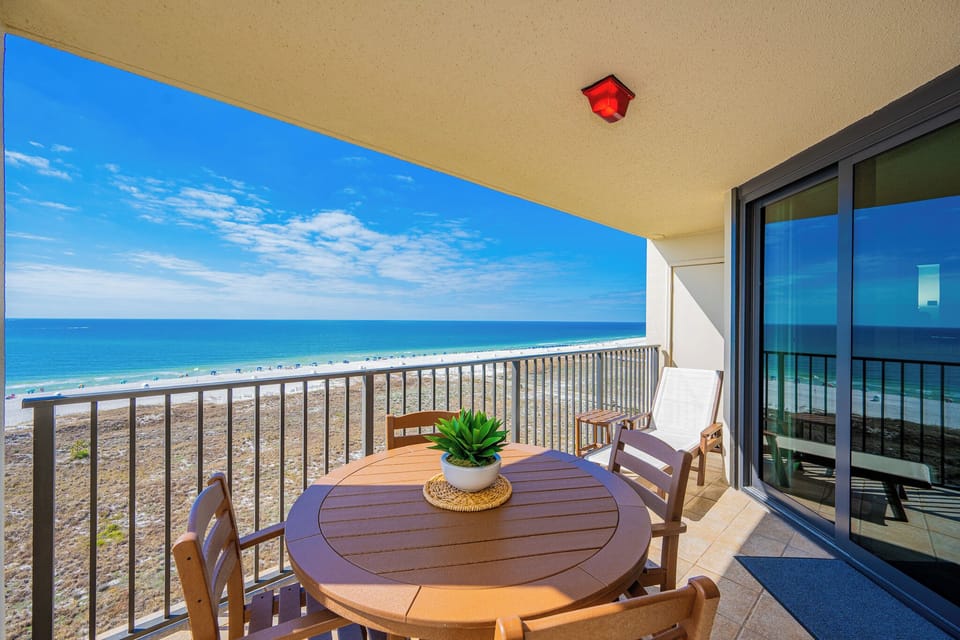 Balcony with a round table and chairs offers a serene view of the beach and turquoise gulf waters under a bright blue sky