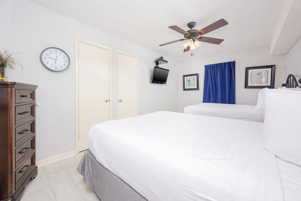 Bright guest bedroom featuring two queen beds, a wall-mounted TV, a wooden dresser, and a blue curtain adding a pop of color
