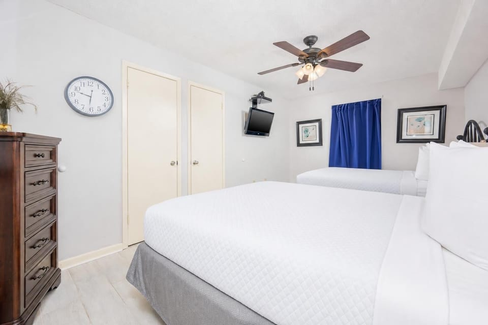 Bright guest bedroom featuring two queen beds, a wall-mounted TV, a wooden dresser, and a blue curtain adding a pop of color