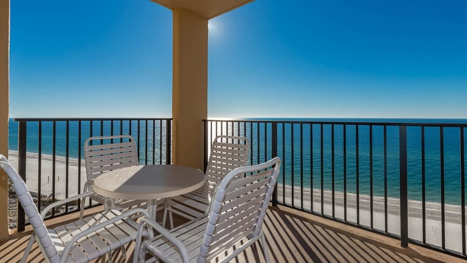 A cozy balcony features a round table and white chairs, offering a view of the tranquil beach and sparkling water