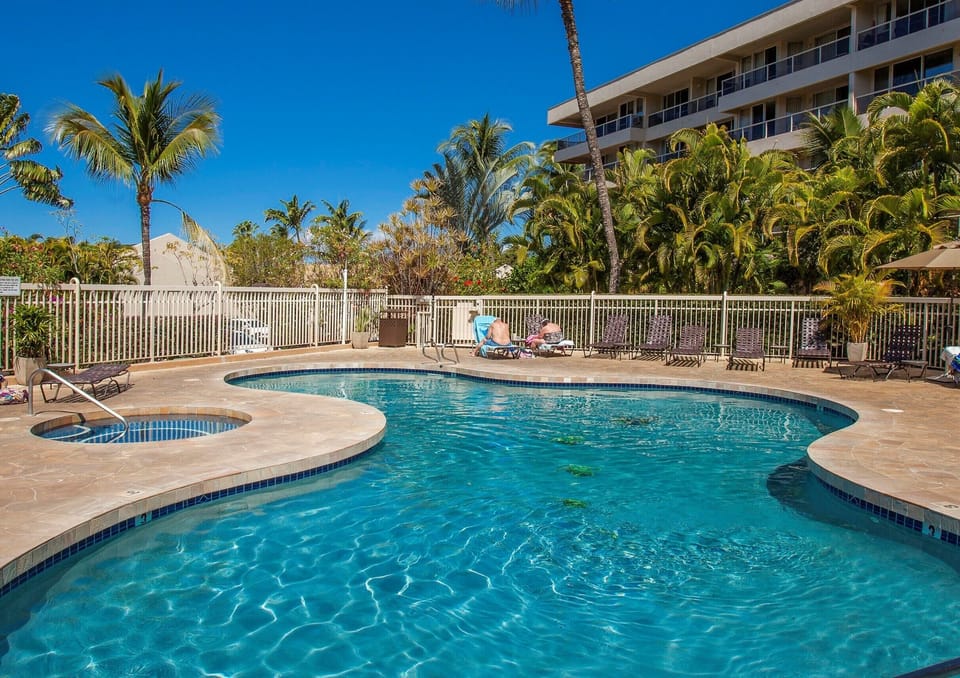 An inviting pool with sparkling blue water awaits, framed by swaying palm trees and tropical greenery. Lounge chairs and a jacuzzi promise relaxation under sunny skies.