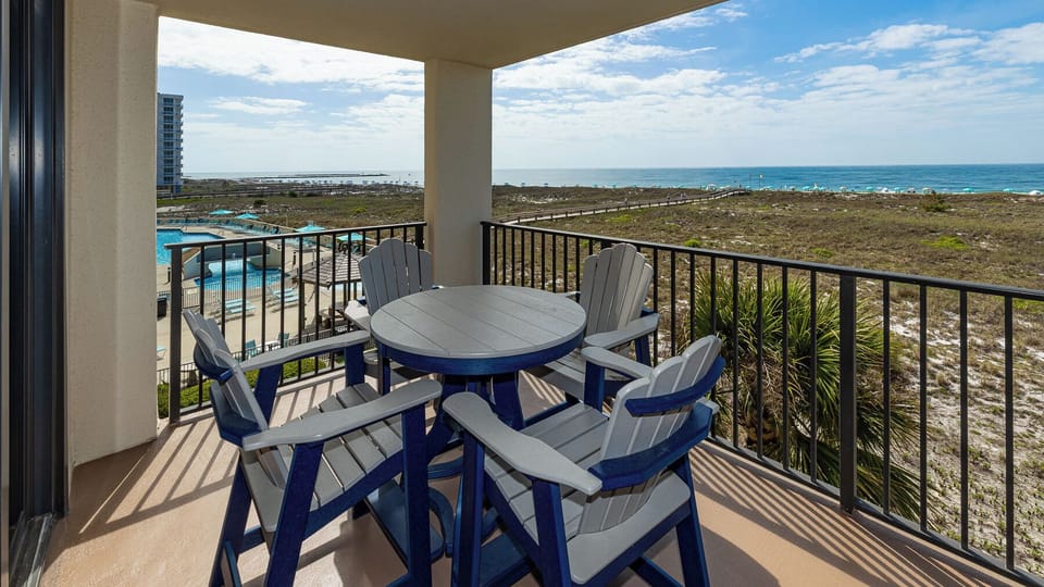 Balcony features a round table and four high-backed chairs, offering a view of the beach and gentle waves beyond