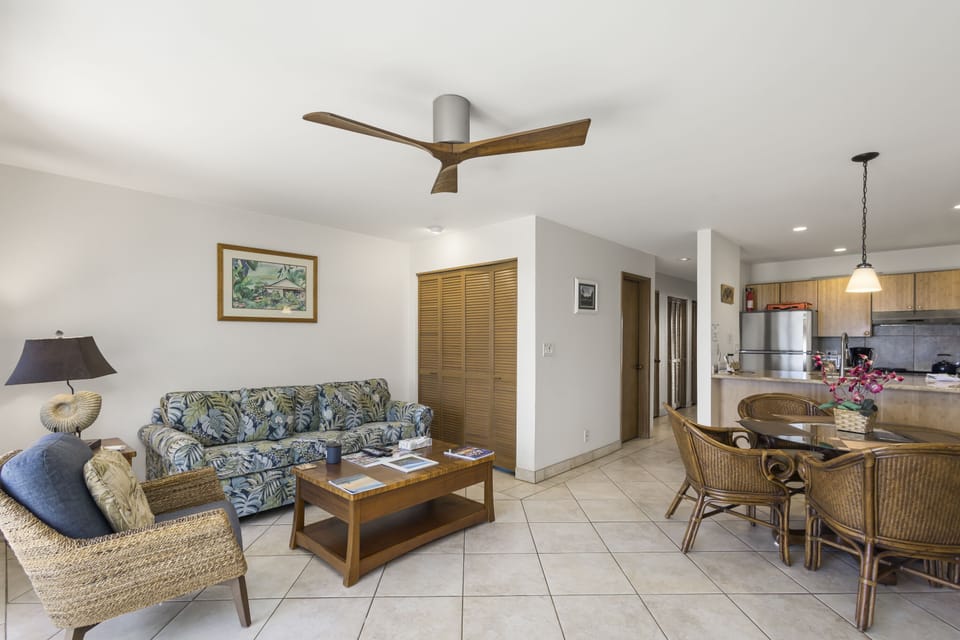 Relax in your open-concept living space featuring a modern ceiling fan, tropical-print sofa, and wicker dining set steps from a fully equipped kitchen.