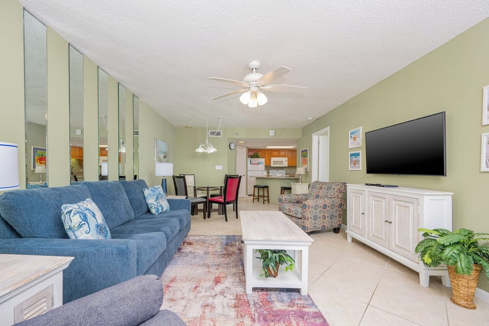 Cozy living area features a blue sofa with decorative pillows, a patterned chair, and a view into the kitchen space