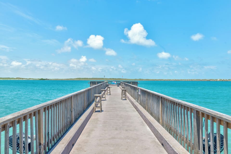 A scenic wooden pier extends into crystal-clear turquoise waters under brilliant blue skies with puffy white clouds.