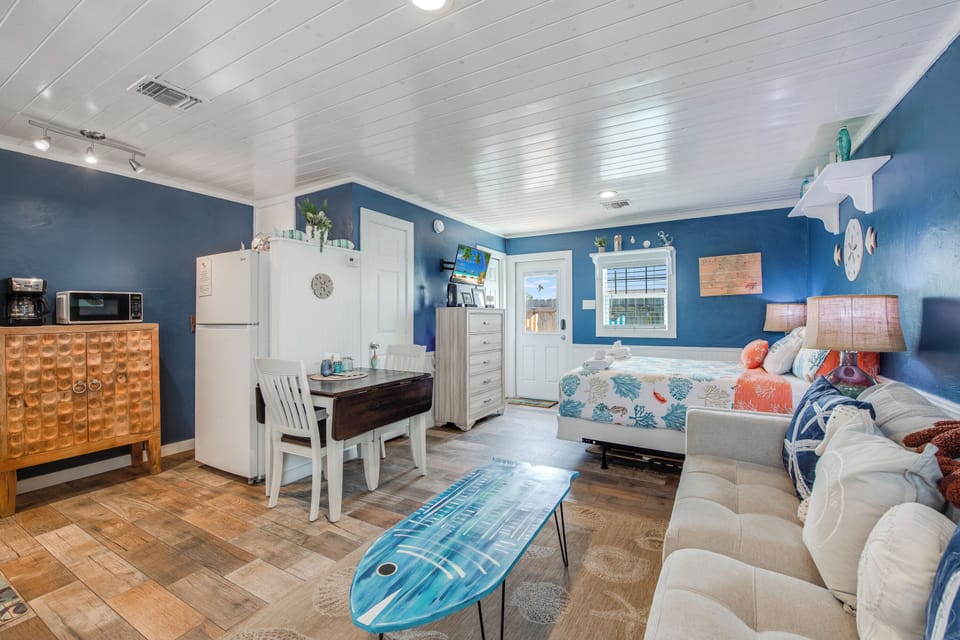 Relax in your coastal-themed studio with vibrant blue walls, full kitchen, and surfboard coffee table for that perfect beach vibe.
