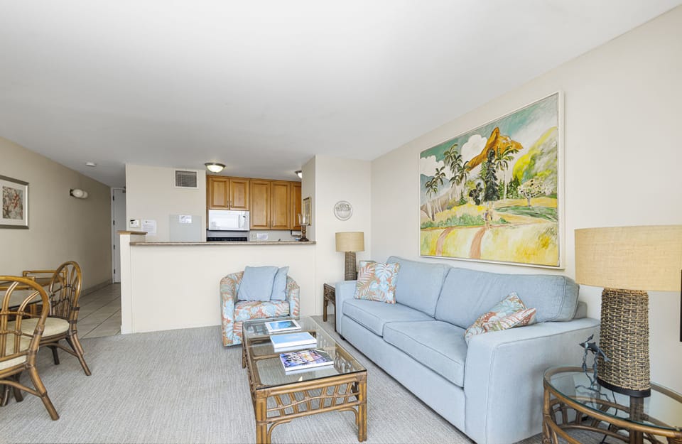 Relax in your airy, open-concept living space featuring a cozy sofa, tropical artwork, and a fully equipped kitchenette just steps away—perfect for unwinding after beach adventures.