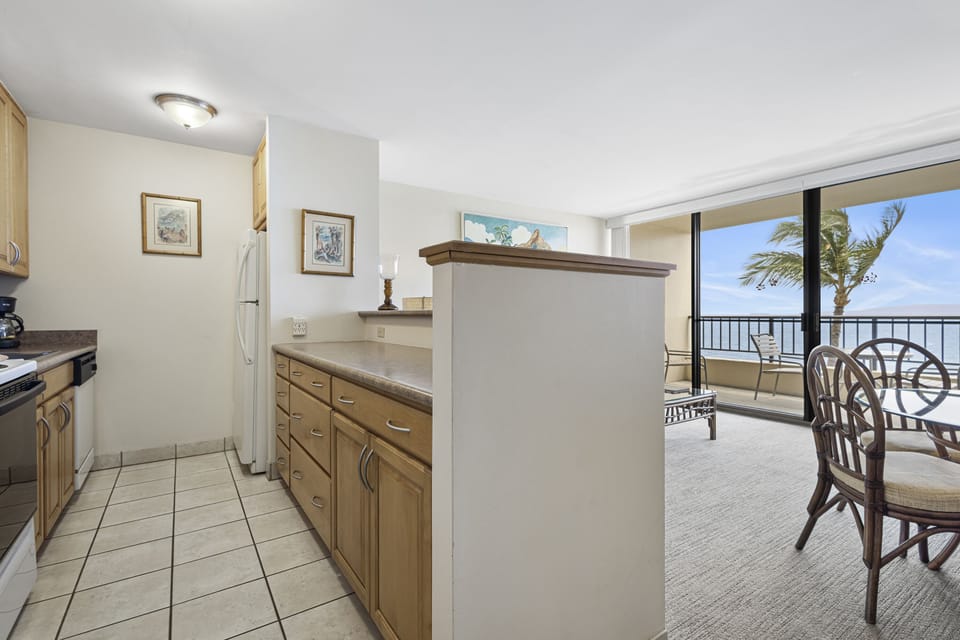 Your fully equipped kitchen flows into the living space, where an ocean view stretches across the horizon, framed by swaying palms and the open balcony beyond.