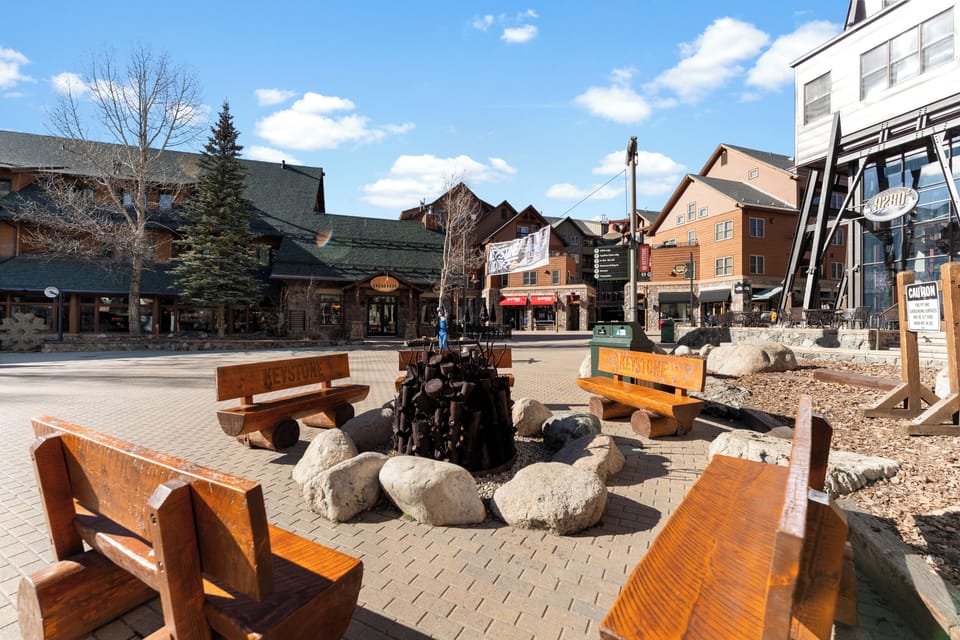 This charming village square features shops, dining, and a central fire pit gathering area, all set against a backdrop of mountain architecture under sunny skies.