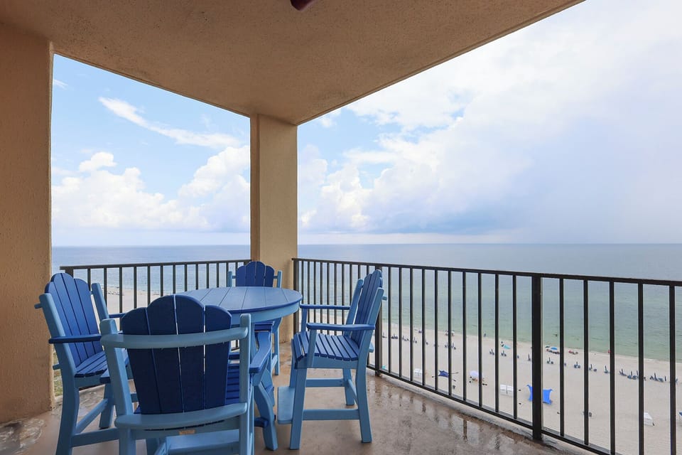 The balcony features a round blue table with matching chairs, offering a serene view of the beach and calm waters below