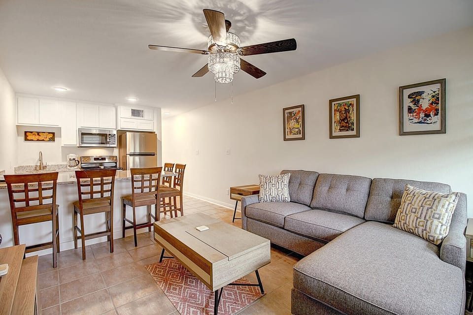 Relax in your open-concept living space with a sparkling chandelier fan, modern sectional, and fully equipped kitchen for comfortable stays.