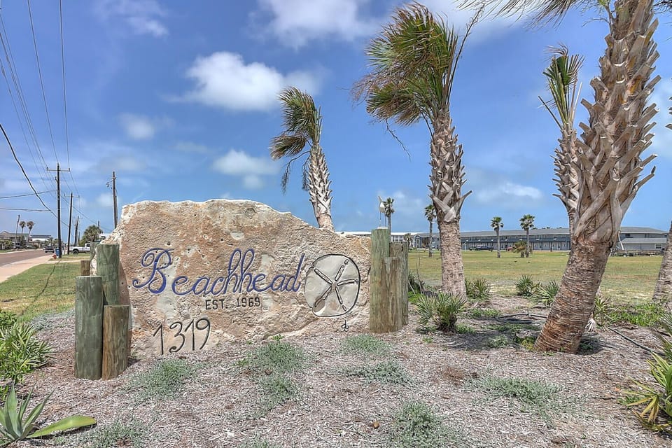 Coastal property entrance featuring decorative stone signage surrounded by palm trees in a beachside community setting.