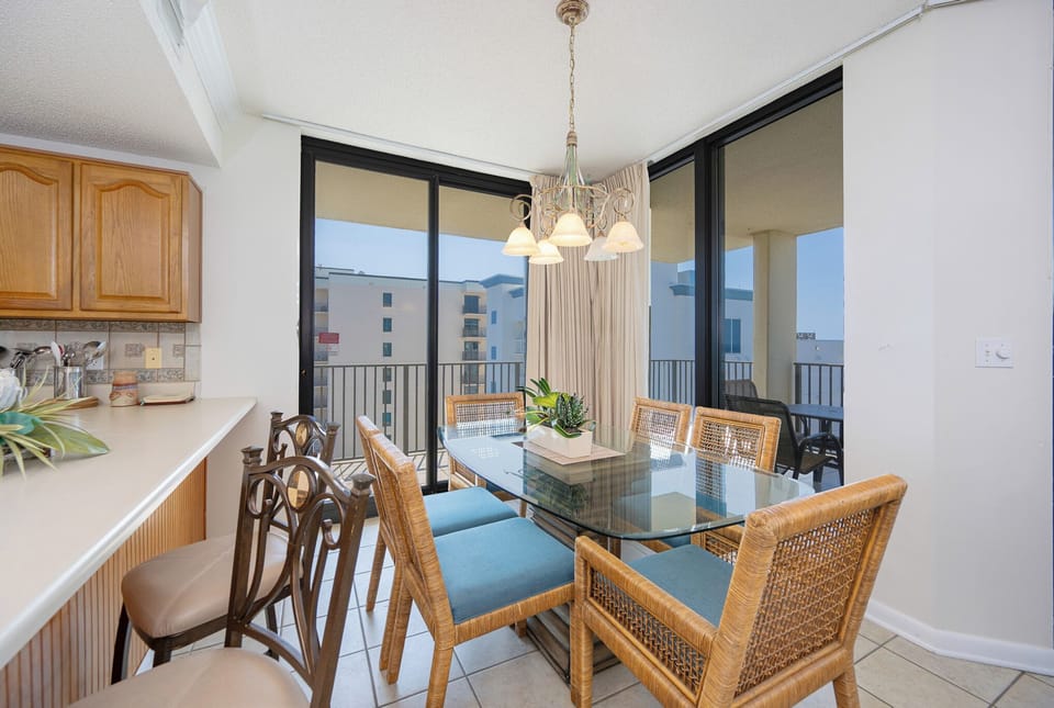 Dining area features a glass table surrounded by wicker chairs, with a view of the beach visible through sliding glass doors