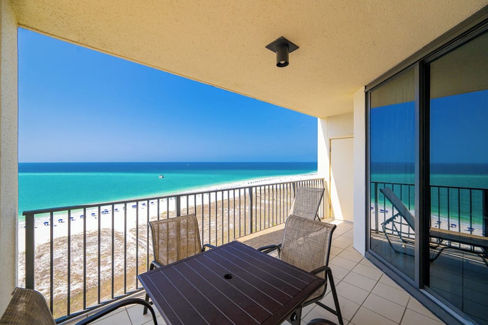 Expansive view of the beach with white sand and gentle waves, framed by lush greenery and a boardwalk leading to the shore