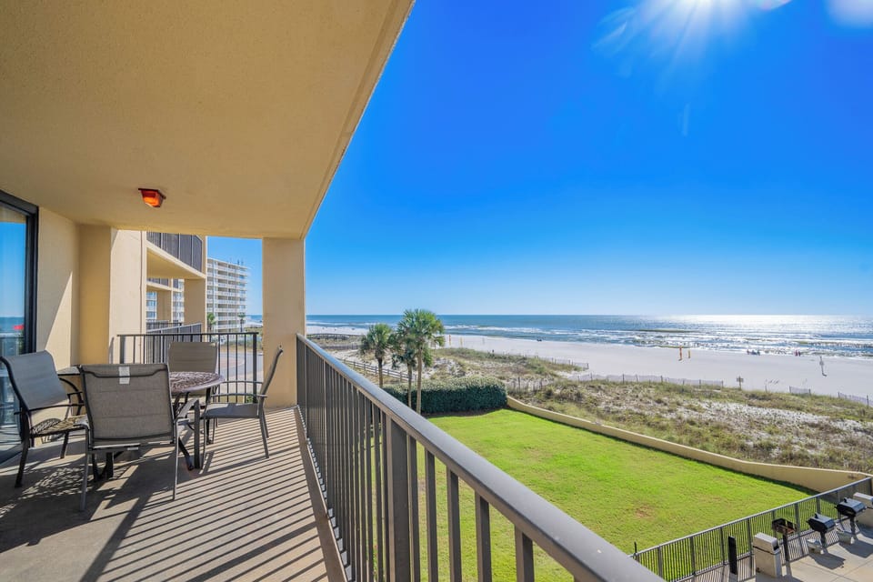 The balcony features a round table with chairs, offering a serene view of the beach and shimmering water under a clear sky