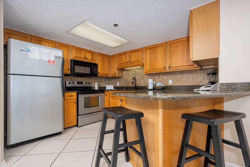 The fully-equipped kitchen features warm wooden cabinetry, a sleek countertop, and a breakfast bar with black stools