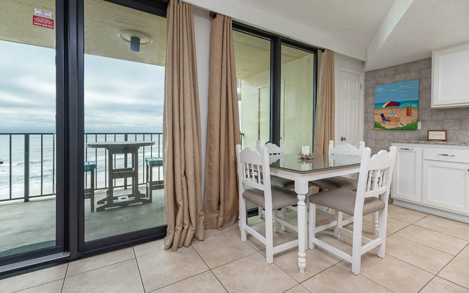 This inviting dining area features a glass-top table, white chairs, and a lovely view of the beach from the balcony