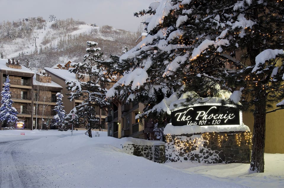 The Phoenix property entrance nestled in a pristine winter wonderland, with ski slopes visible in the background and twinkling lights creating magical mountain ambiance.