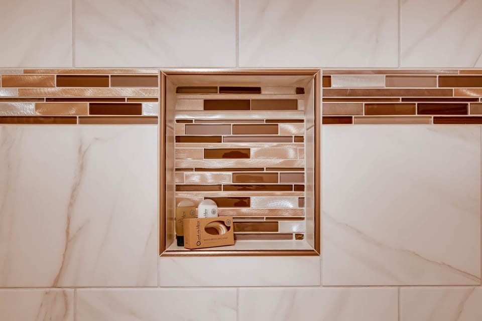 The en suite bathroom has gorgeous white tile, with gold and brown decorative tile lining a shelf in the bathtub surround.