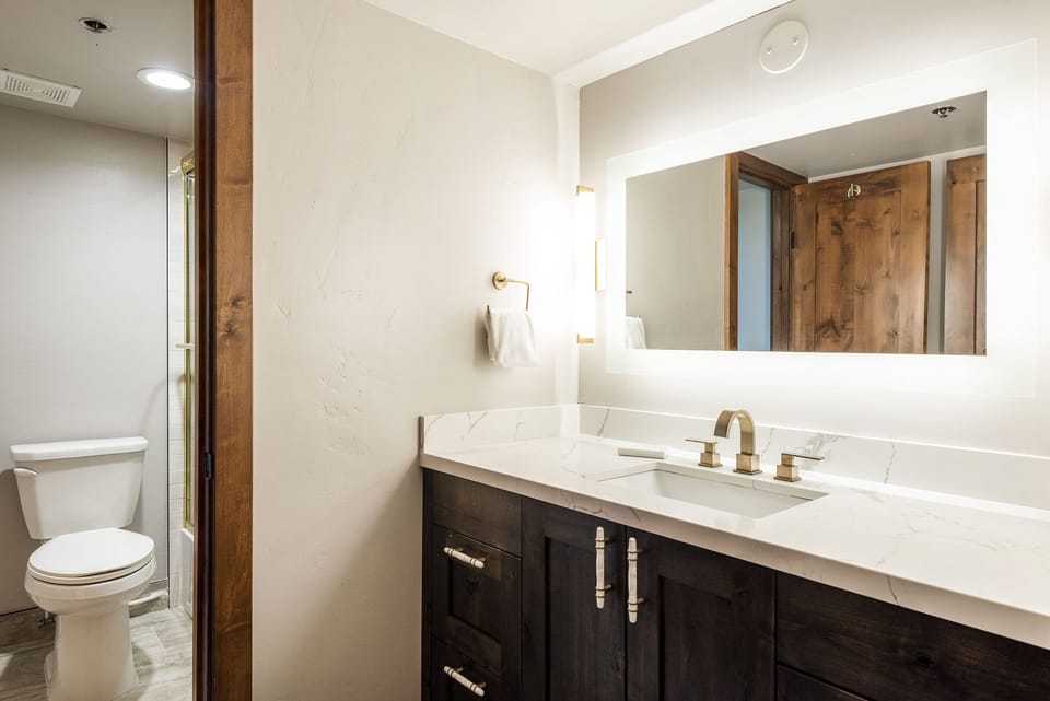 Unit 229 hallway bathroom with dark wood vanity, white countertop and large illuminated mirror.