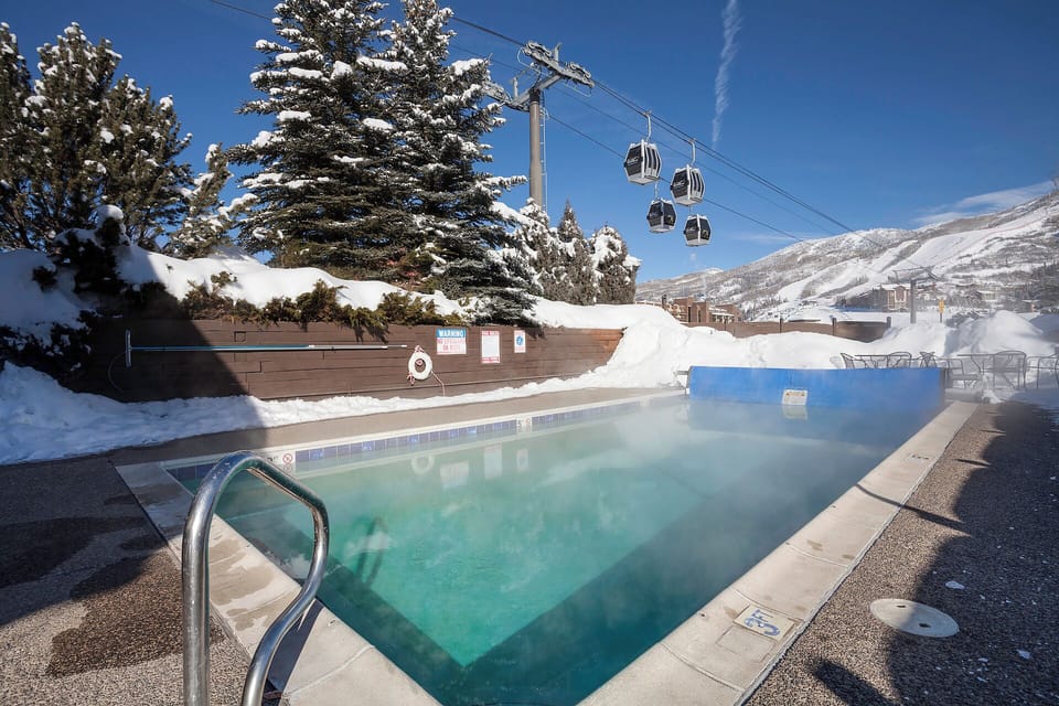 Outdoor heated pool surrounded by snow-covered mountains and evergreen trees, with ski gondolas overhead creating a perfect alpine winter escape.