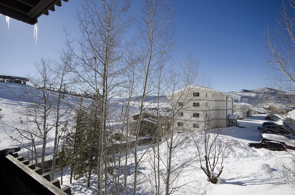 Snow-covered alpine landscape with mountain village buildings visible beyond winter trees and clear blue skies.