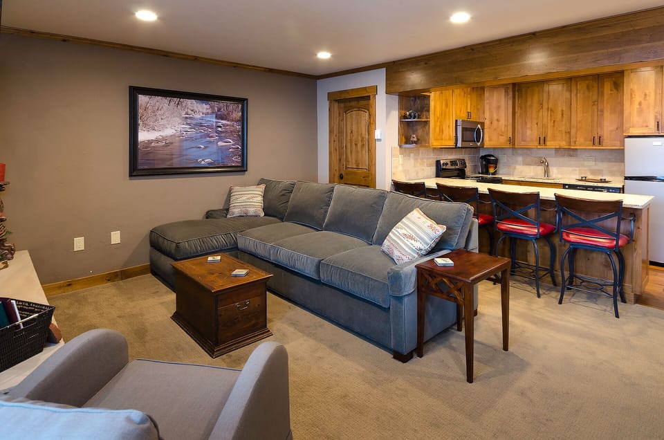 Relax in your open-concept living space featuring a plush sectional sofa, rustic kitchen with warm wood cabinets, and mountain artwork creating the perfect alpine retreat.