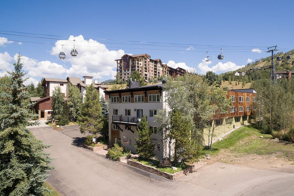 Mountain resort village with ski gondola access and multi-story lodges nestled among evergreen trees under clear blue skies.