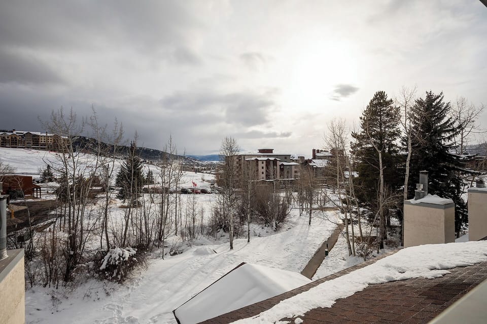 Winter mountain landscape with snow-covered neighborhood buildings and evergreen trees under cloudy skies.