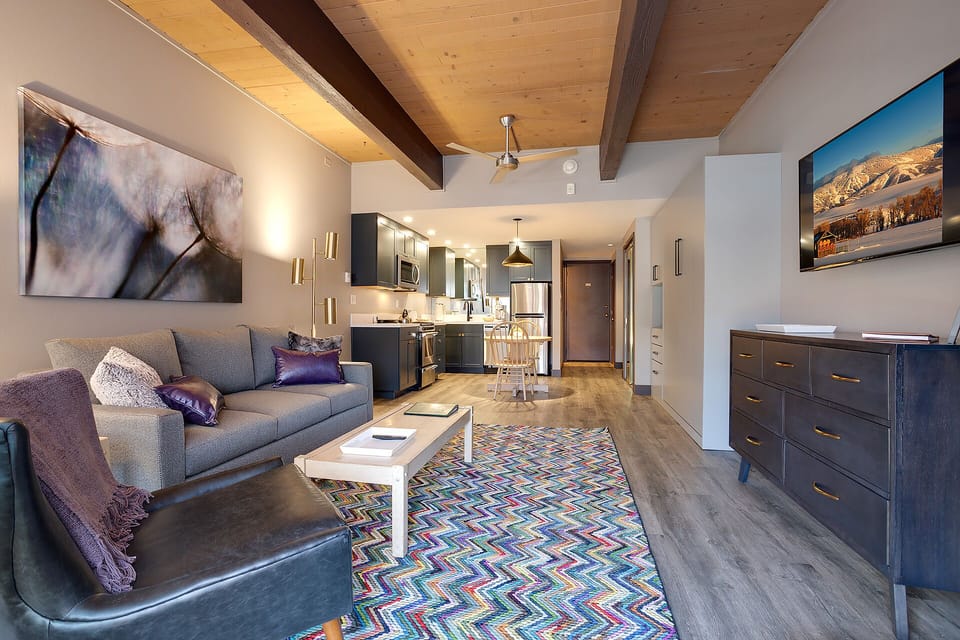 Relax in your stylish open-concept living space featuring exposed wood beams, a cozy sofa, and a fully equipped kitchen.