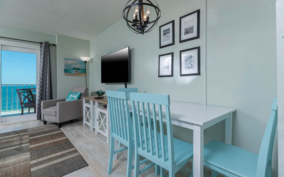 This inviting living area features a large TV, light blue dining chairs, and a stunning beachfront view from the balcony