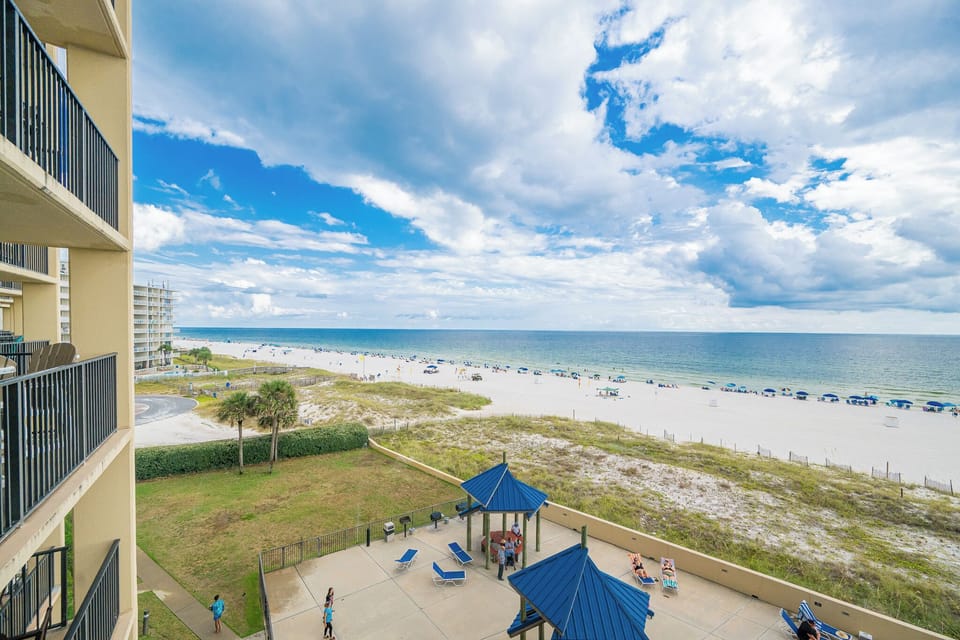 The view showcases a vibrant beach with umbrellas and loungers, framed by lush greenery and a charming pool area below