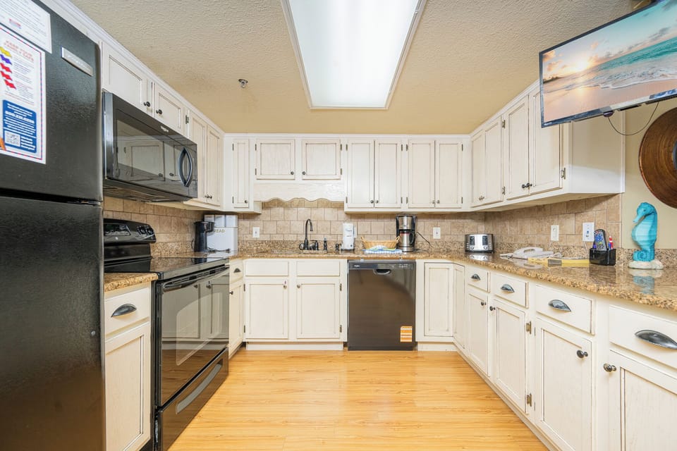 Bright kitchen with white cabinetry, granite countertops, and black appliances, featuring a TV, Keurig and coffee maker