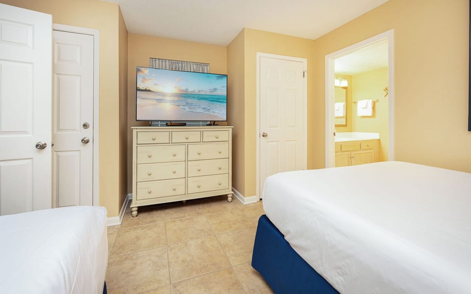 Cozy guest bedroom featuring a large TV, two beds, dresser, and a view into the bathroom with a bright, inviting atmosphere