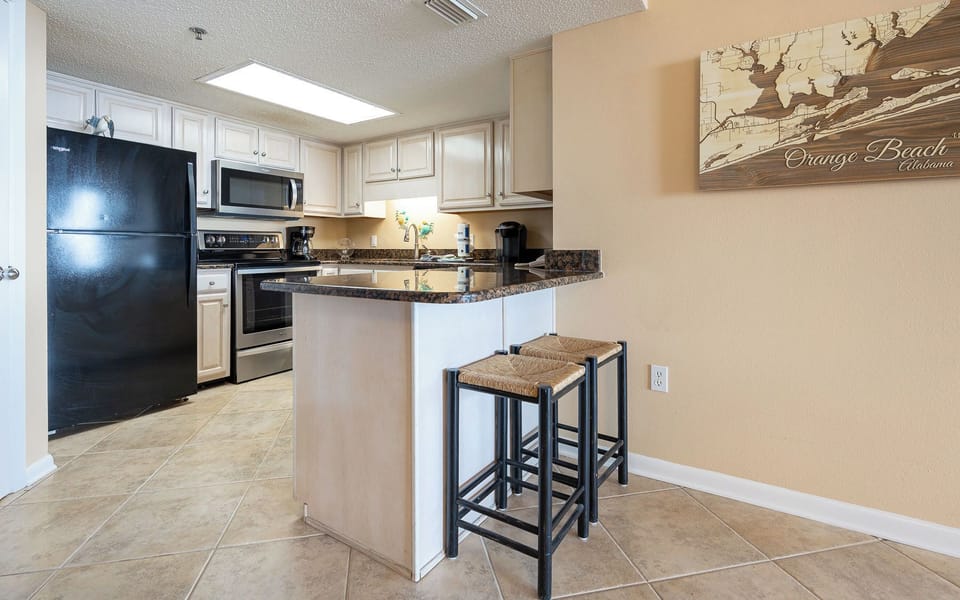 Modern kitchen features dark granite countertops, black appliances, and a breakfast bar with two woven stools