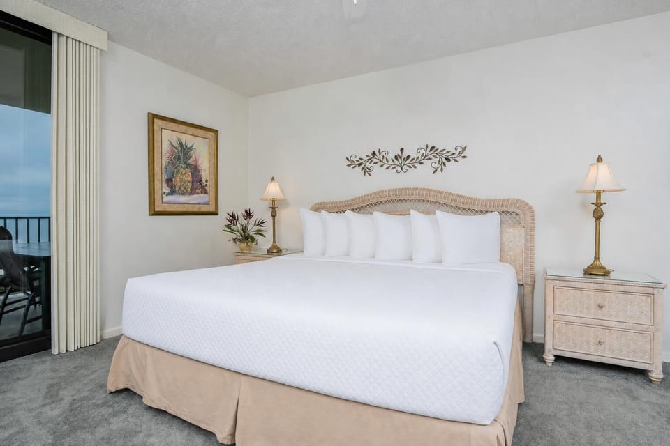 Cozy master bedroom featuring a king bed with plush pillows, a decorative lamp, and a balcony view of the beach beyond